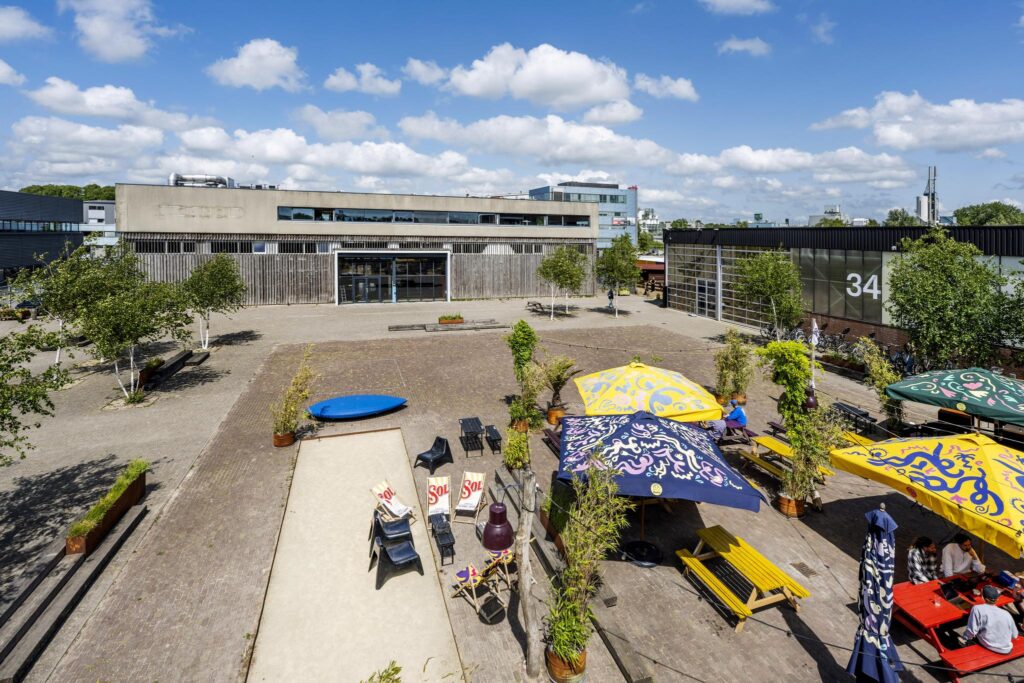 Outdoor seating area with colorful umbrellas and picnic tables in front of creative industrial buildings on Johan van Hasseltweg.